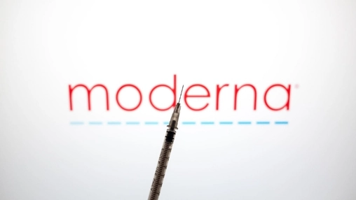Medical syringe in sharp focus against a blurred Moderna logo on a plain white background with blue dashes