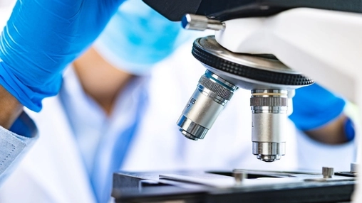 Gloved hands adjusting the objective lenses of a laboratory microscope, white lab coats visible behind