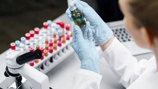A gloved scientist examines a small amber vial beside a rack of colour-capped test tubes and a microscope